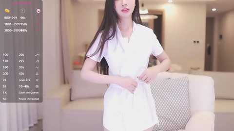 Media: Video of an East Asian woman with long black hair, wearing a white dress, standing in a modern, brightly lit living room with white furniture.