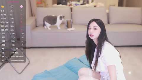 Media: Video of an Asian woman with long black hair, wearing a white shirt, sitting on a blue mat, playing a video game. Background includes a grey sofa, a white cat, and a TV screen displaying game stats.