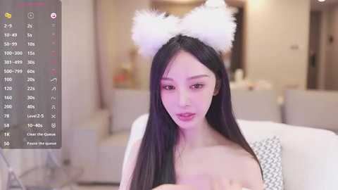 Media: A video of an East Asian woman with long black hair, wearing white cat ears, and a white top, sitting on a couch in a modern, softly-lit room.