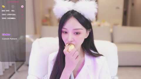 Media: Video of an East Asian woman with long black hair, wearing white cat ears and a white coat, eating a donut in a modern living room.