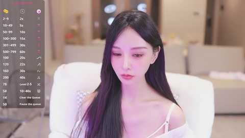 Media: Video of an East Asian woman with long black hair, wearing a white off-shoulder top, seated indoors. Background features a blurred, modern living room with beige furniture.