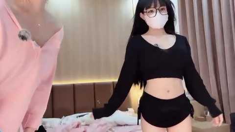 Media: Video of an Asian woman with long black hair, wearing a black crop top and shorts, holding hands with a man in a pink shirt. Background shows a modern bedroom with a brown headboard and soft lighting.