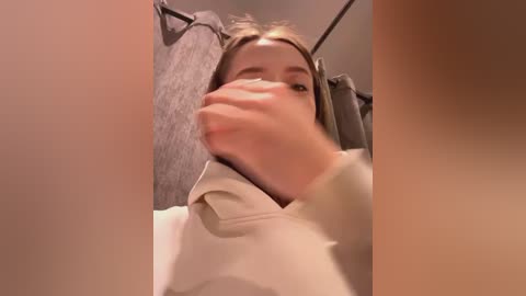 Media: Video of a woman with light skin and straight, shoulder-length brown hair, wearing a white hoodie, partially covering her face with her right hand, standing in a bathroom with grey towels and beige walls.