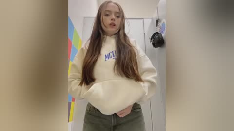 Media: Video of a young Caucasian woman with long, straight brown hair, wearing a cream hoodie with \"MC\" logo and green pants, standing in a brightly lit, colorful hallway.