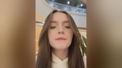 Media: Video of a young woman with long, straight brown hair, fair skin, and light makeup, wearing a white top. Background features a modern, brightly lit room with curved walls and soft lighting.