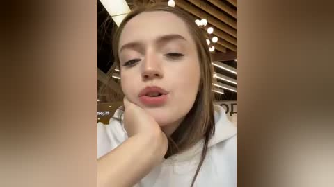 Media: Video of a young woman with pale skin and long brown hair, wearing a white top, resting her chin on her hand, in a modern, brightly lit caf\u00e9 with wooden ceiling beams and large windows.