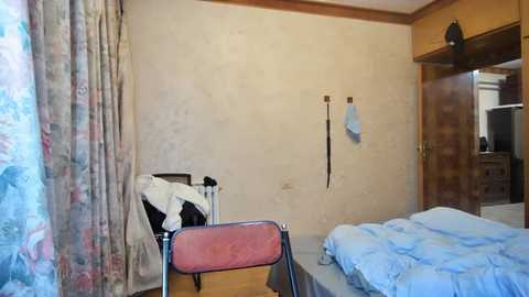 Media: Video of a small, cluttered bedroom with beige walls, floral curtains, a metal-framed bed, and a chair. Clothes are strewn about, and a wooden door leads to another room.