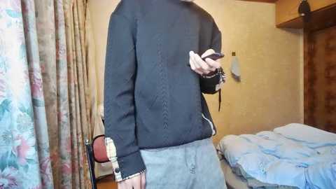 Media: A video of a person wearing a dark sweater, gray shorts, and a wristwatch, holding a smartphone. Background includes a bed with blue sheets, floral curtains, and a wooden door.