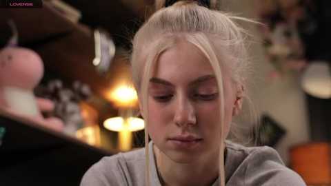 Media: Video of a young woman with platinum blonde hair in a bun, wearing a gray top, focused on her makeup in a softly lit, cozy room with a blurred background.