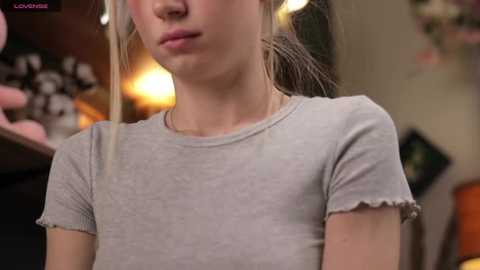 Media: Video of a young Caucasian woman with fair skin, wearing a light gray, short-sleeved top, with her long blonde hair tied back. The background is blurred, showing a warm, cozy room with soft lighting.