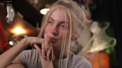 Media: Video of a young woman with light blonde hair in a messy bun, wearing a grey T-shirt, blowing a bubble. Background is blurry with warm lighting and red decor.