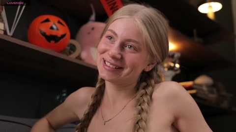 Media: Video of a smiling, topless young woman with blonde braided hair, wearing a gold necklace, in a dimly lit room with Halloween decorations.