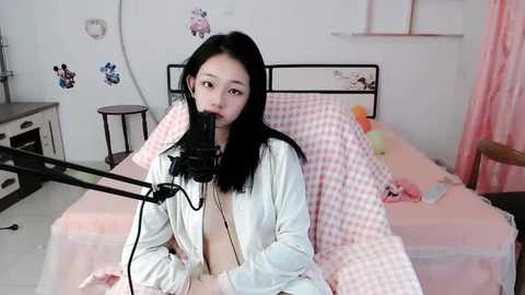 Media: A video of an East Asian woman with long black hair, wearing a white robe, sitting on a bed with pink checkered sheets, singing into a microphone in a cozy bedroom with pastel decor.