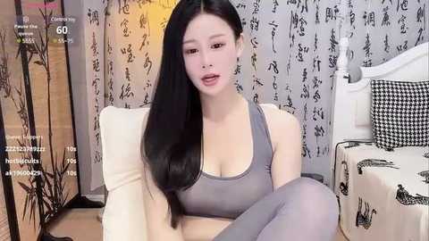 Media: A video of an East Asian woman with long black hair, fair skin, and a slender physique, wearing a gray sports bra and leggings. She sits on a white couch in a room with a black-and-white Japanese-themed backdrop.