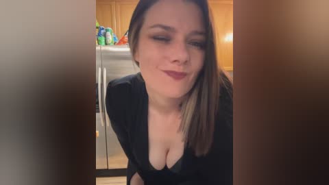 Media: Video of a smiling, light-skinned woman with shoulder-length brown hair, wearing a black top with a deep V-neckline, leaning forward in a kitchen with wooden cabinets and a colorful fruit bowl.