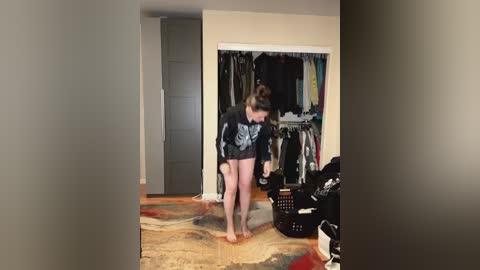 Media: Video of a woman with short brown hair wearing a black graphic T-shirt and black shorts, standing barefoot on a patterned rug in a cluttered bedroom with an open closet filled with clothes and a laundry basket.