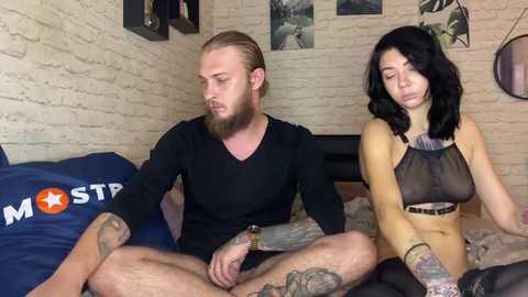 Media: A video of a bearded man in a black long-sleeve shirt, and a tattooed woman in a sheer black top, sitting on a bed with blue and white pillows.