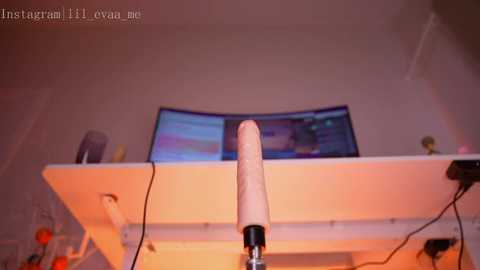 Media: Video of a realistic, flesh-colored dildo mounted on a white desk, with a computer monitor displaying a colorful image in the background. The room is dimly lit, creating a warm ambiance.