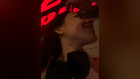 Media: Video of a joyful woman with long dark hair, wearing a black top, smiling widely while a hand gently touches her face, in a dimly lit, red-lit room.