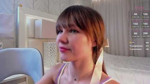 Media: Video of a young Caucasian woman with light skin, brown hair, and brown eyes, wearing a white tank top, sitting indoors, with a virtual reality headset on.