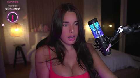 Media: Video of a curvy, brunette woman with large breasts wearing a red tank top, singing into a blue-lit microphone in a dimly lit bedroom.