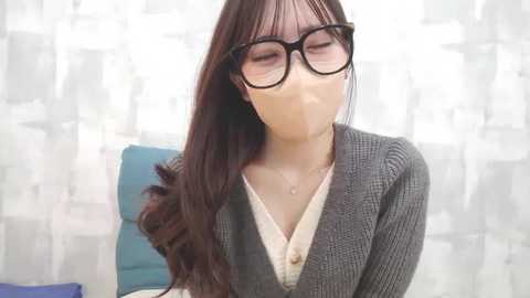 Media: Video of a young Asian woman with long dark hair, wearing large black-framed glasses, a beige blouse, and a grey cardigan, with a face mask covering her mouth and nose. She is adjusting her hair in a cozy, white-patterned room.