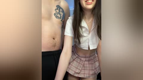 Media: Video of a smiling young woman with long brown hair and a small tattoo on her shoulder, wearing a white cropped top and a plaid skirt. Behind her, a shirtless man with a large tattoo on his chest.