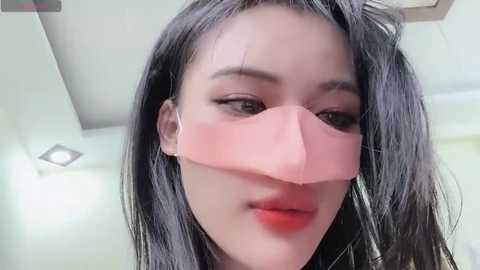 Media: Video of a young Asian woman with long, straight black hair, wearing a pink surgical mask, red lipstick, and subtle makeup, in a bright, modern room with white walls and a ceiling light.