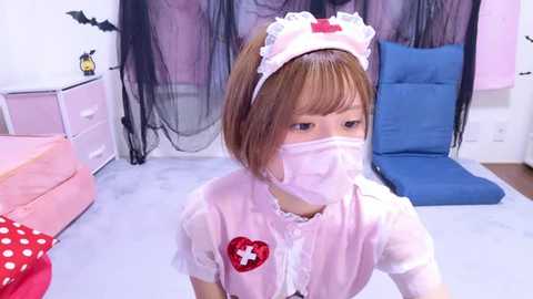 Media: Video of a young Japanese girl with straight brown hair, wearing a pink nurse outfit, including a headband and mask, in a pastel-themed room with pink furniture and a blue cushion.