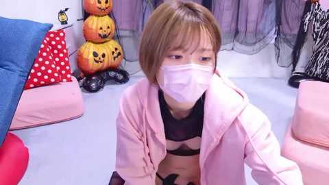 Media: Video of an Asian woman in a pink jacket, black bra, and mask, sitting in a room with Halloween decorations: pumpkins, polka-dotted pillow, and zebra print.