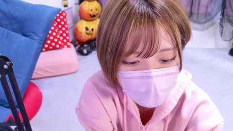 Media: A video of an Asian woman in a pink hoodie and mask, sitting on a bed with Halloween decorations and stuffed toys in a colorful room.