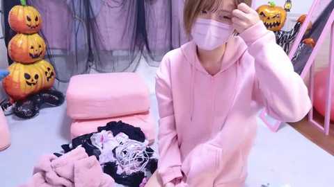 Media: Video of a young Asian woman in a pink hoodie, mask, and glasses, sitting on a bed with Halloween-themed decorations, including pumpkin lights and a black net.