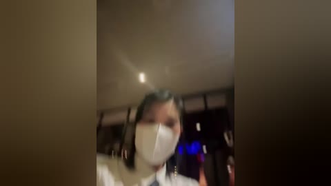 Media: Video of a person wearing a white mask, standing in a dimly lit room with blurred background. The person's face is partially obscured by the mask, and they appear to be looking forward.