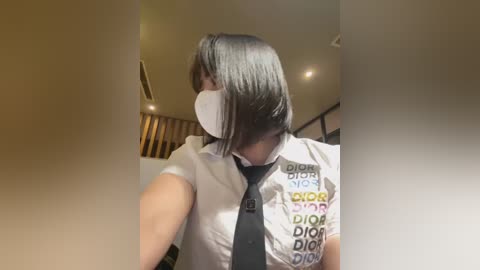 Media: Video of an Asian woman with straight black hair, wearing a white blouse with a black tie and a white surgical mask, standing indoors with beige walls and a wooden ceiling.