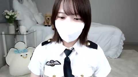 Media: Video of a young Asian woman in a white military uniform with a black tie, wearing a white mask, seated on a white rug in a minimalistic bedroom with a bed and stuffed toys.