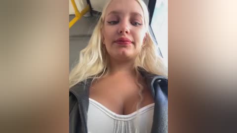 Media: Video of a blonde woman with fair skin, wearing a gray jacket over a white tank top, sitting on a bus. Background shows bus interior with yellow handles and blue window.