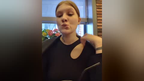 Media: Video of a young woman with fair skin and straight, light brown hair, wearing a black top with a cutout. She puckers her lips, standing indoors near a window with flowers.