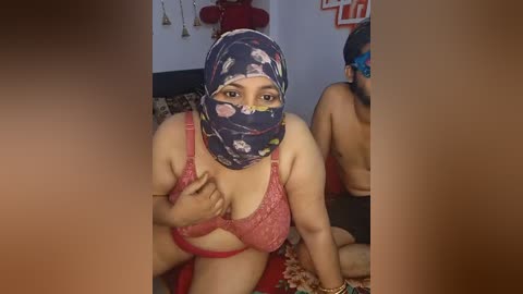 Media: Video of a plus-sized woman with light brown skin, wearing a floral-patterned headscarf, red bra, and a colorful beaded necklace, sitting on a couch in a dimly lit room.