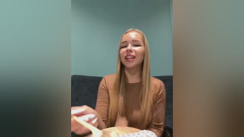 Media: Video of a smiling young woman with long blonde hair, wearing a brown top, holding a spoon and eating noodles in a grey couch setting against a teal wall.