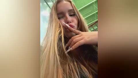 Media: Video of a young blonde woman with long hair, wearing a black top, blowing a kiss with her left hand, in a green-walled room with metal bars.