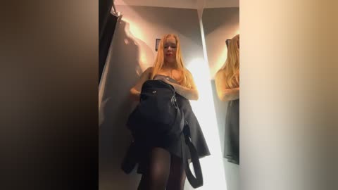 Media: Video of a young, slender, blonde woman with long hair, wearing a black dress and holding a black backpack, walking down a dimly lit hallway with beige walls and mirrors reflecting her.