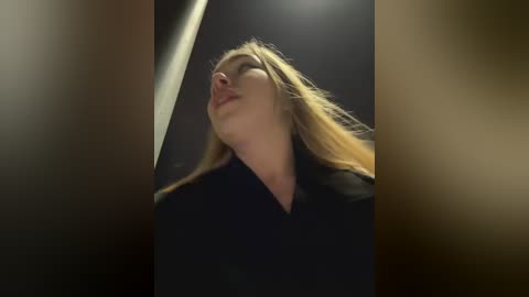 Media: Video of a woman with long blonde hair, wearing a black coat, looking upward with a serene expression. The background is dark and blurred, emphasizing her silhouette.