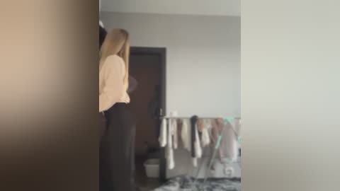 Media: Video of a blonde woman in a beige blouse and black pants standing in a dimly lit room with a closet door open, displaying hanging clothes.