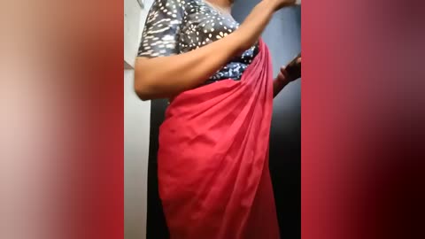 Media: Video of a woman with medium brown skin wearing a dark blouse with white polka dots and a bright red saree, standing in a dimly lit room with red and gray walls.