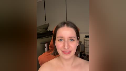 Media: Video of a young, fair-skinned woman with straight, shoulder-length brown hair, wearing only a towel, smiling slightly. She sits on a bed with a beige headboard in a dimly lit room.