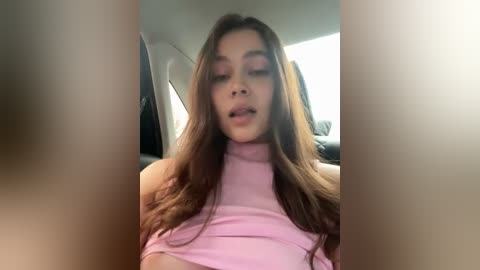 Media: A blurry video of a young woman with long, wavy brown hair, wearing a light pink, off-the-shoulder crop top, seated in a car. Her expression is neutral, with slightly parted lips. The background is out of focus, showing a car interior.