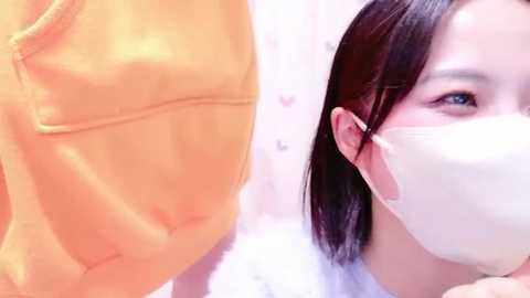 Media: A video of an Asian woman with straight black hair, wearing a white surgical mask, looking to the right, while a person in an orange hoodie is partially visible on the left.