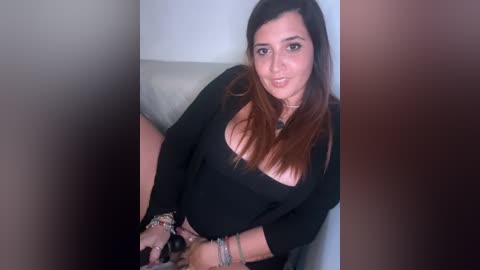 Media: Video of a smiling Latina woman with long brown hair, wearing a black top, sitting on a beige couch. She has a curvy figure and wears multiple bracelets.