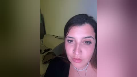 Media: Video of a young woman with light skin and dark hair, wearing a black top, standing in a dimly lit room. She has a nose ring, and her makeup is subtle with dark eyeliner.