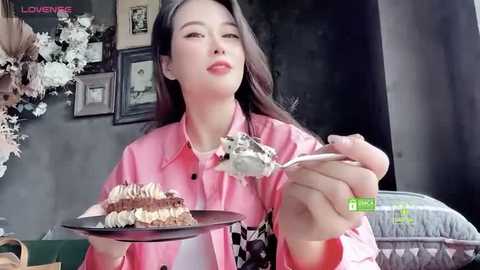 Media: Video of an East Asian woman with long black hair, wearing a pink blouse, eating cake with a fork, surrounded by framed pictures and floral decorations.